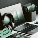 Elegant Workspace with Ticketing Elements: A photo of a sleek, minimalist workspace with a high-end laptop, a pair of stylish headphones, and event tickets in green and black hues. The workspace is organized on a white table, providing a clean and modern look that subtly hints at the profitability of ticket reselling.
